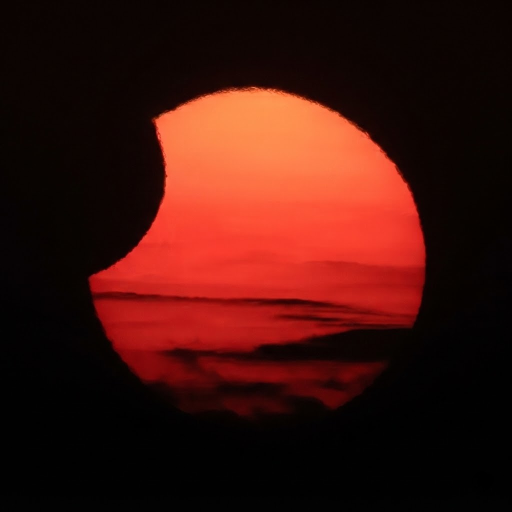 A close-up shot of a partial solar eclipse against a deep red and orange twilight sky with soft, distant clouds.